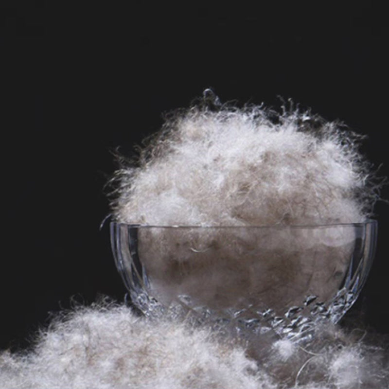 Natural down feathers overflowing a clear glass bowl against a dark background.