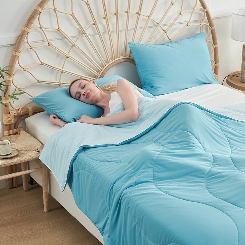 Woman sleeping peacefully beneath a light blue APSMILE cool blanket on a bed with matching pillowcases, set against a rattan headboard.