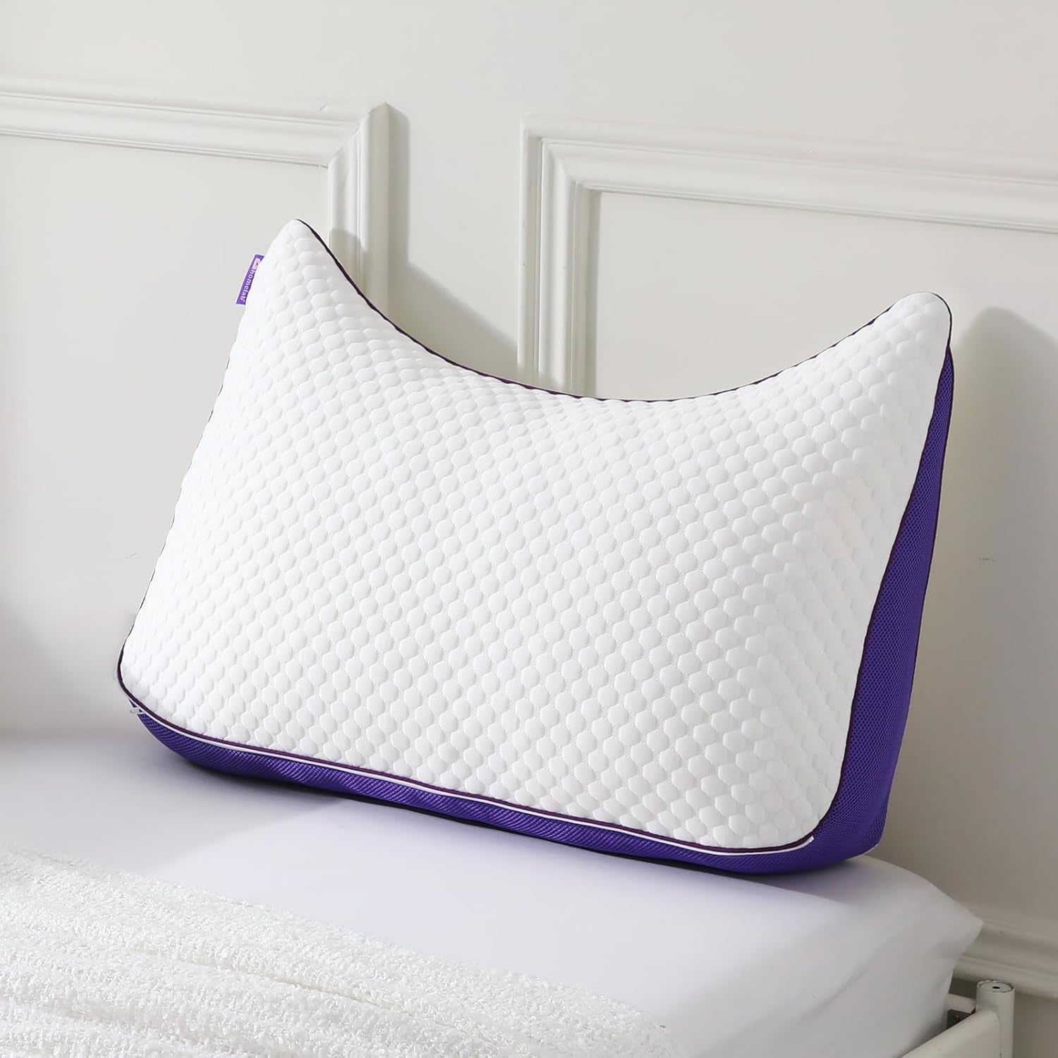 Close-up of an APSMILE crescent-shaped pillow with a white, textured surface and purple trim on a white bed.