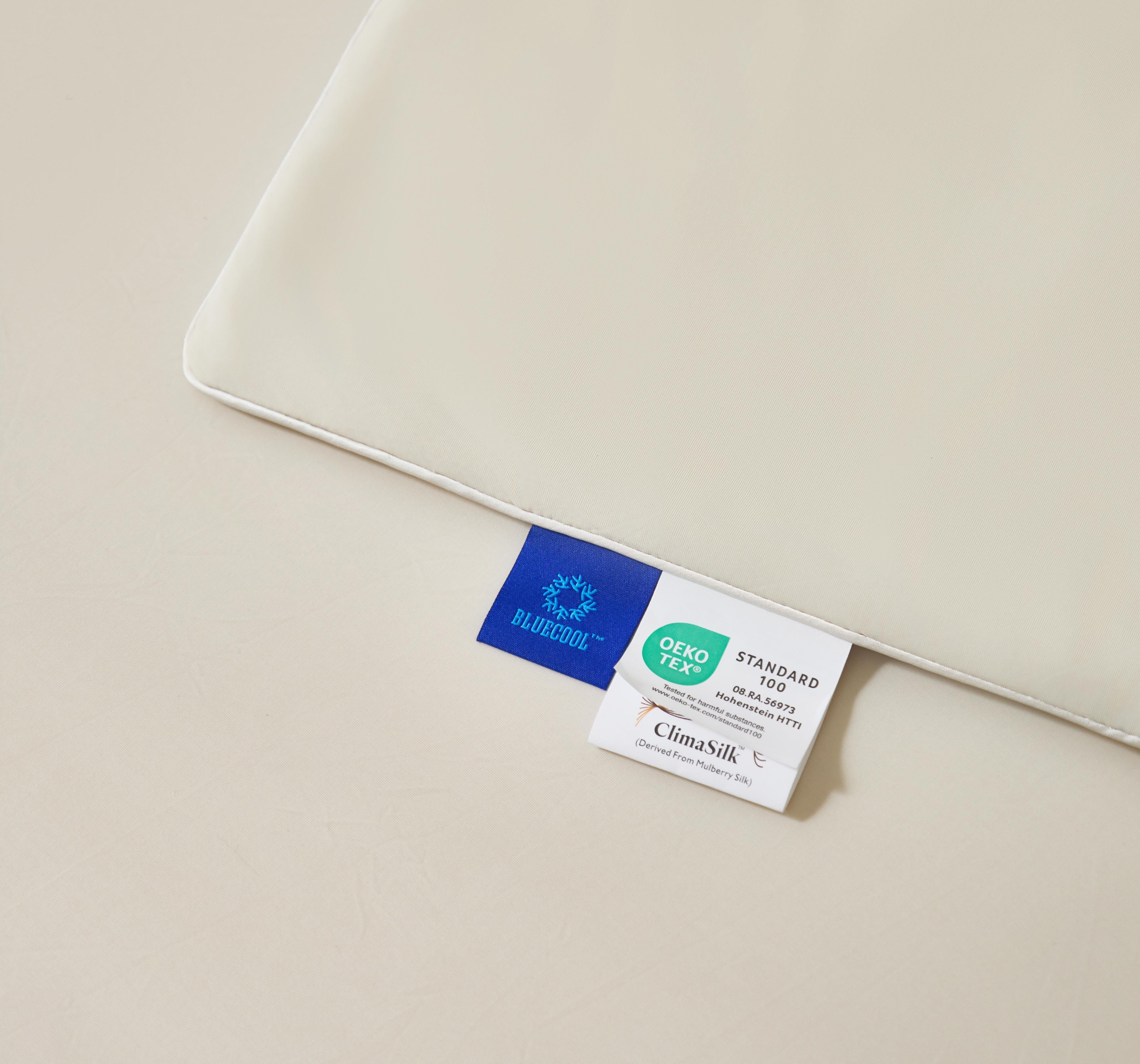 Close-up of a beige pillowcase corner with two fabric tags indicating Bluecool and Oeko-Tex Standard 100 certifications, plus ClimaSilk derived from Mulberry Silk.