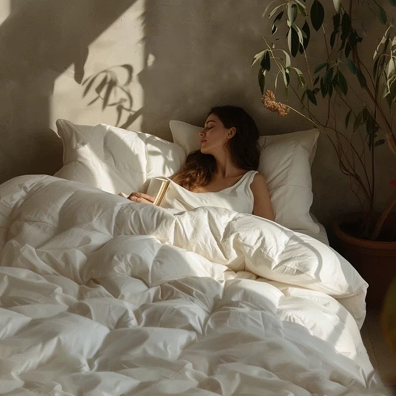 Woman in white tank top relaxing on a bed covered with APSMILE white down comforter, holding a book, with her eyes closed.