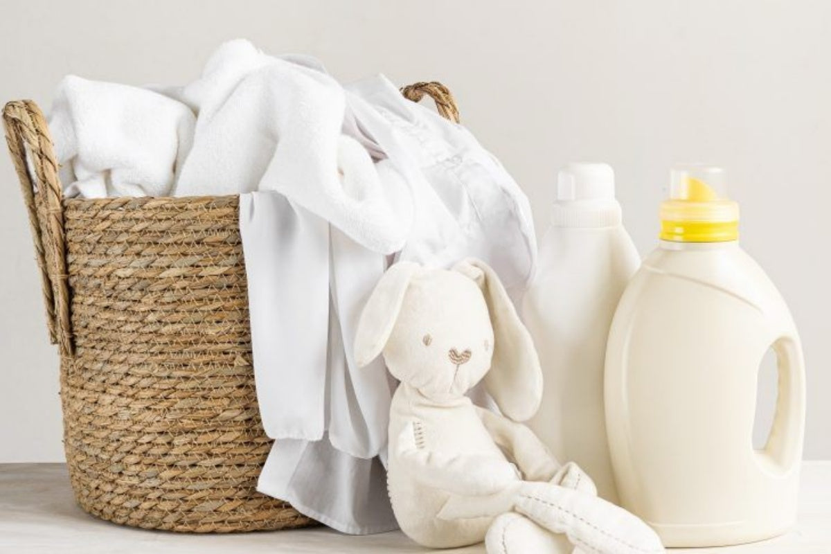 A woven basket overflowing with freshly laundered white clothes is positioned beside a stuffed bunny toy and three bottles of laundry detergent against a light background.