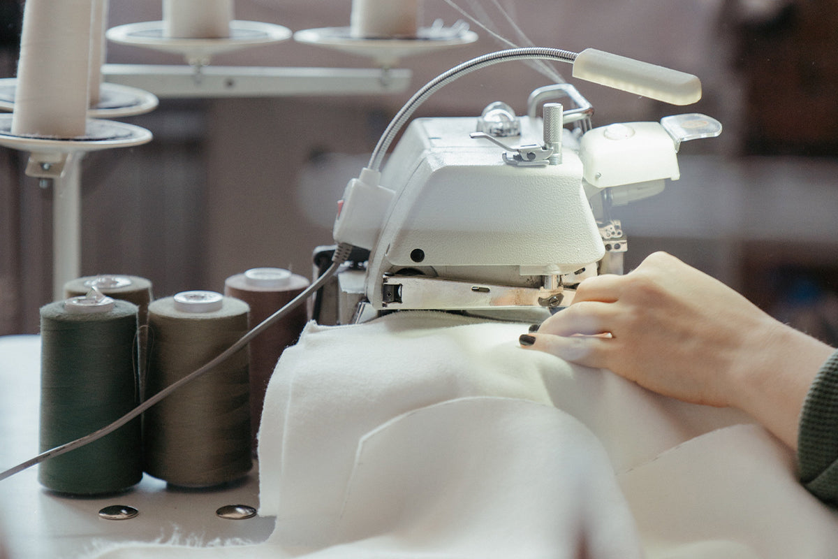 Close-up of hands guiding APSmile duvet through a white sewing machine equipped with a lamp; spools of olive green, brown, and off-white thread stand to the side.