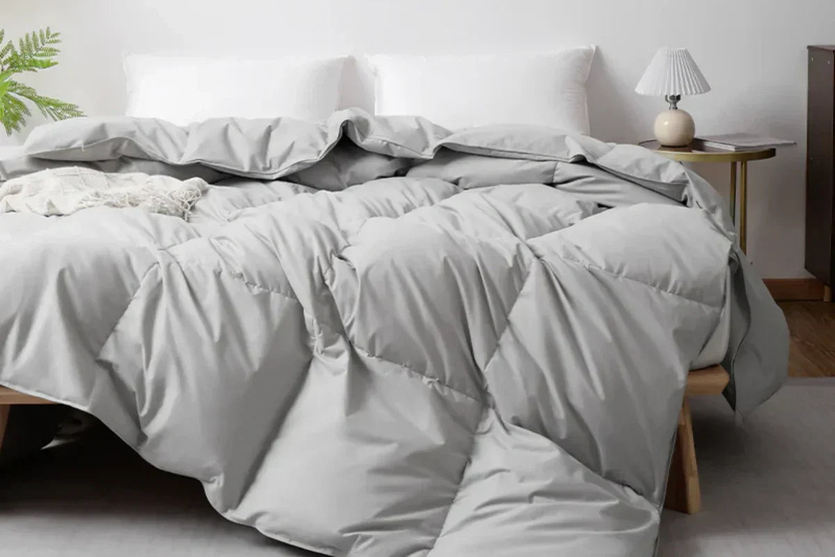 Bedroom setting with a light gray APSmile duvet covering a bed, accented by white pillows and a side table with a lamp and a book.