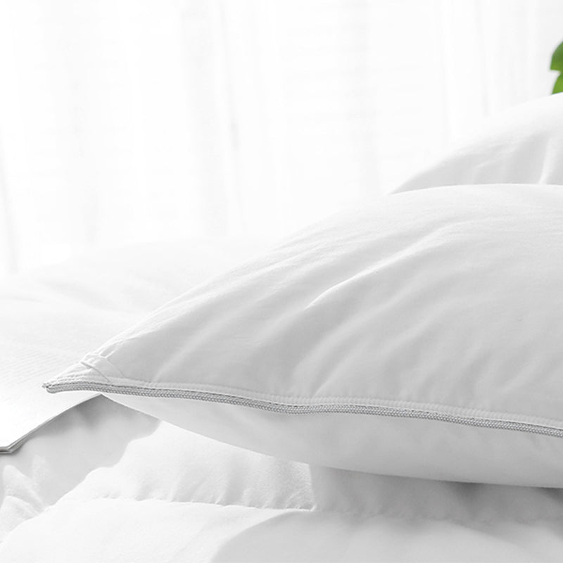 Close-up of a white pillow with a Silver stitched border laying on a white bed.