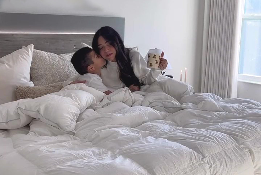 A mother and child are lying in bed with an APSMILE down comforter. The child is turned towards the mother and is kissing her cheek. The mother is holding a teddy bear mug. The bed has white linens by AP SMILE. Tall, thin candles are at the side of the bed. A window is visible in the background.  